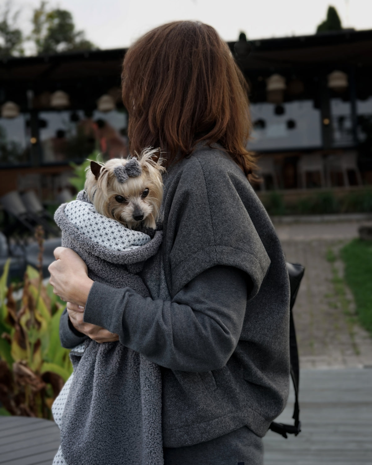 Woman carrying a small cream dog with a grey bouclé hair bow, wrapped in a grey blanket outdoors