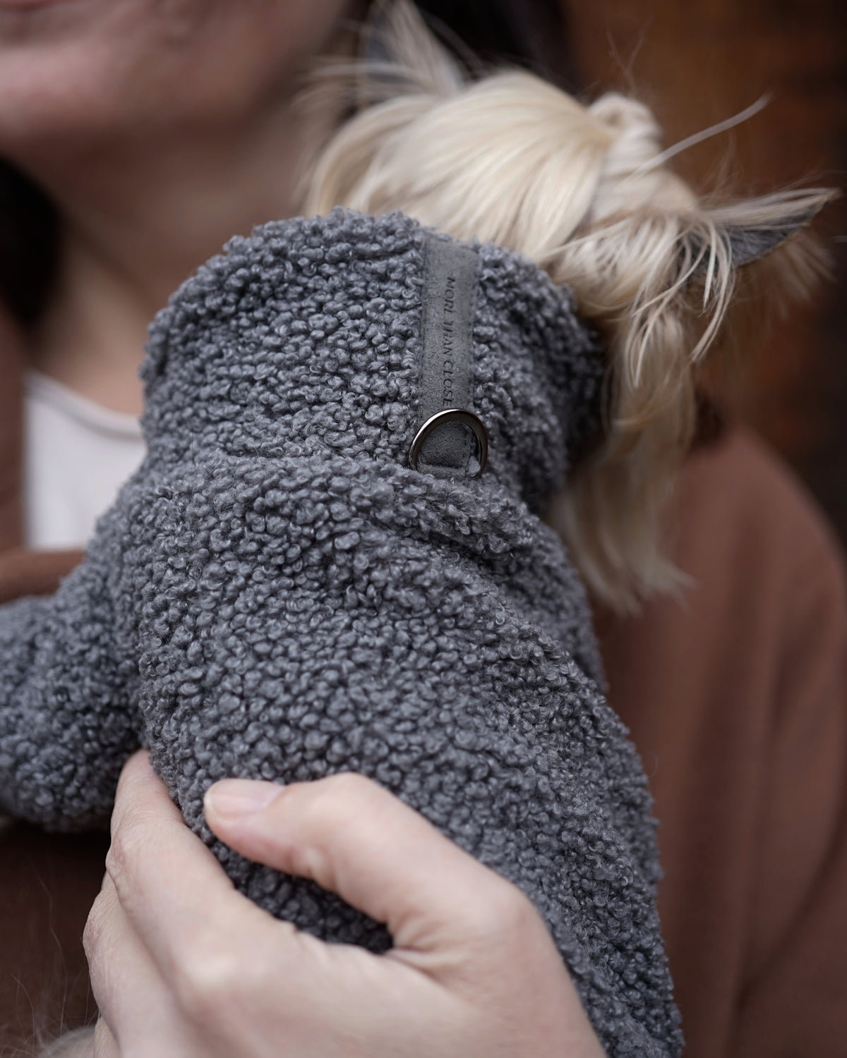 Close-up of a grey bouclé pullover as a small black dog is carried, the suede tab and metal ring visible at the collar