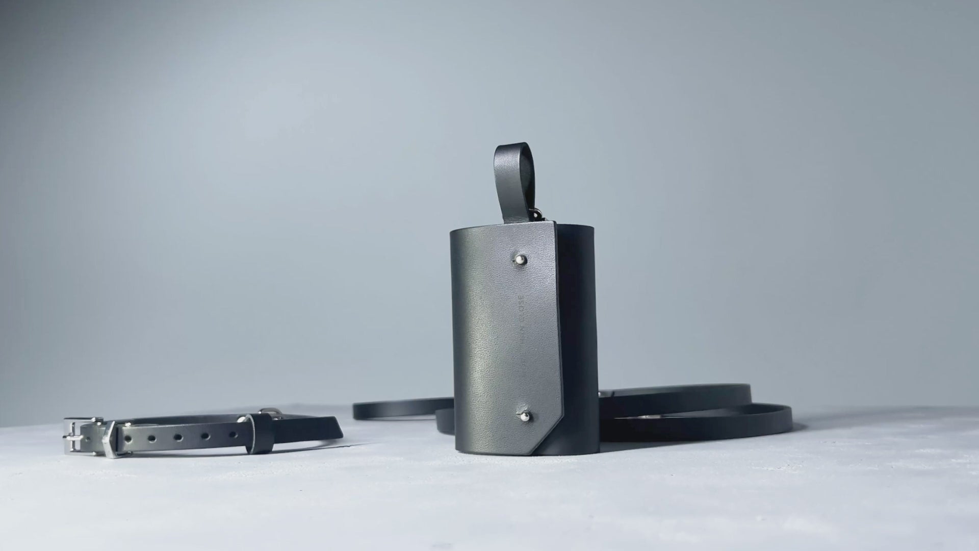 Black small dog walking set with collar, leash and waste bag holder displayed on concrete against a dusty blue background.